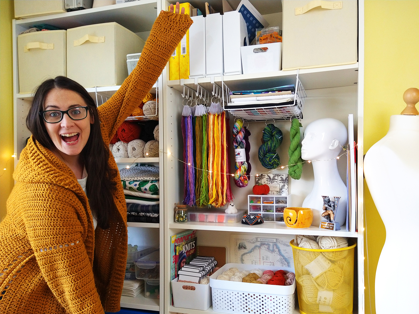 Michelle is standing in front of two white Ikea bookcases full of colourful and inspiring craft supplies like embroidery floss, yarn and fabric. She's wearing a mustard yellow crocheted cardigan and looking very proud of the whole setup!